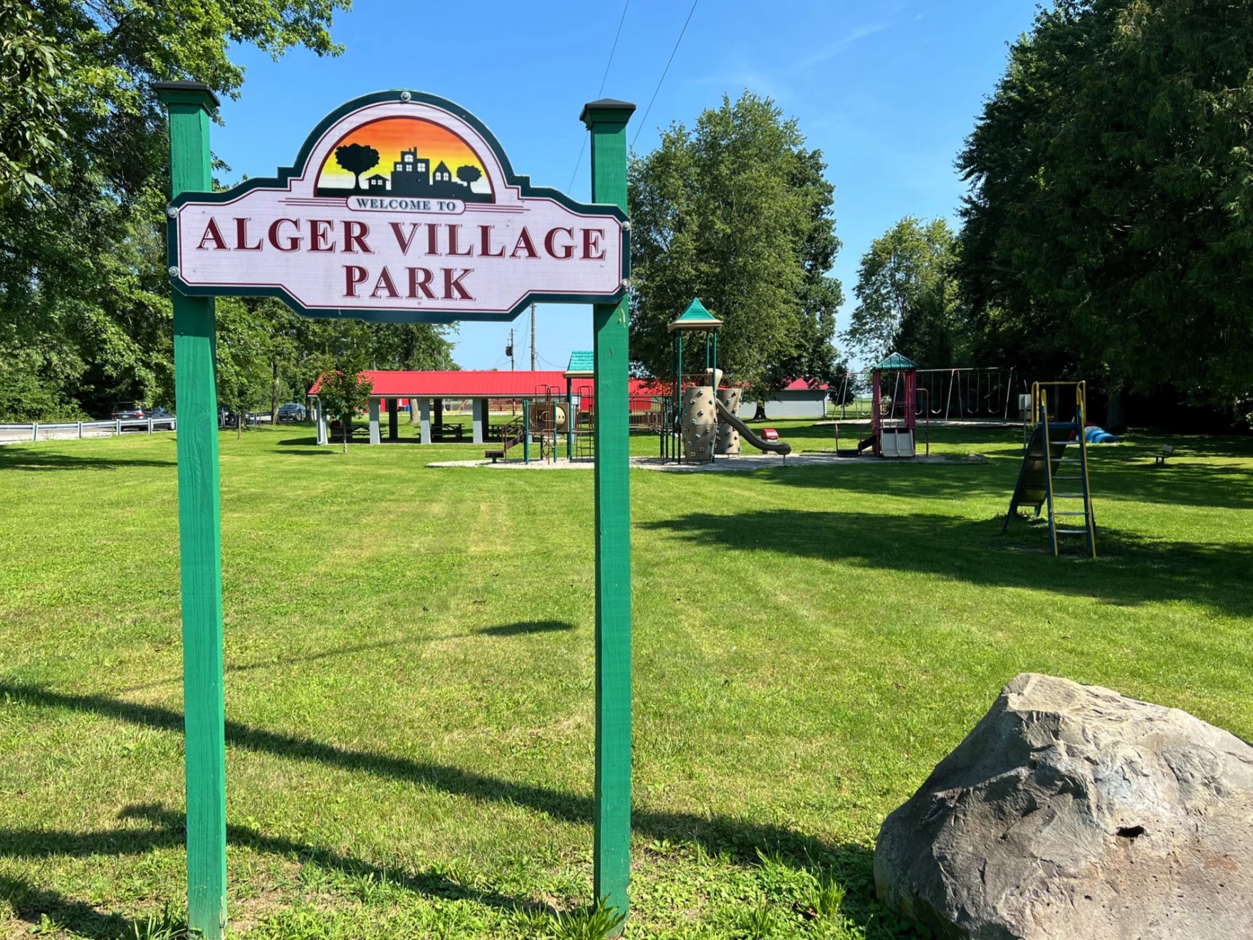 Alger Village Park entrance sign with playground and pavilion in the background.
